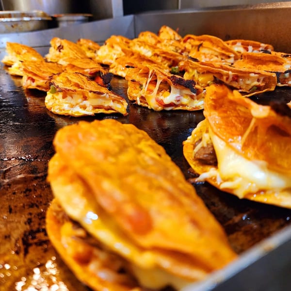Numerous cheesy birria tacos sizzle on a dark, greasy griddle at Flying Burro.