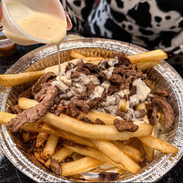 Creamy white sauce being poured over a foil container of golden french fries and steak pieces.